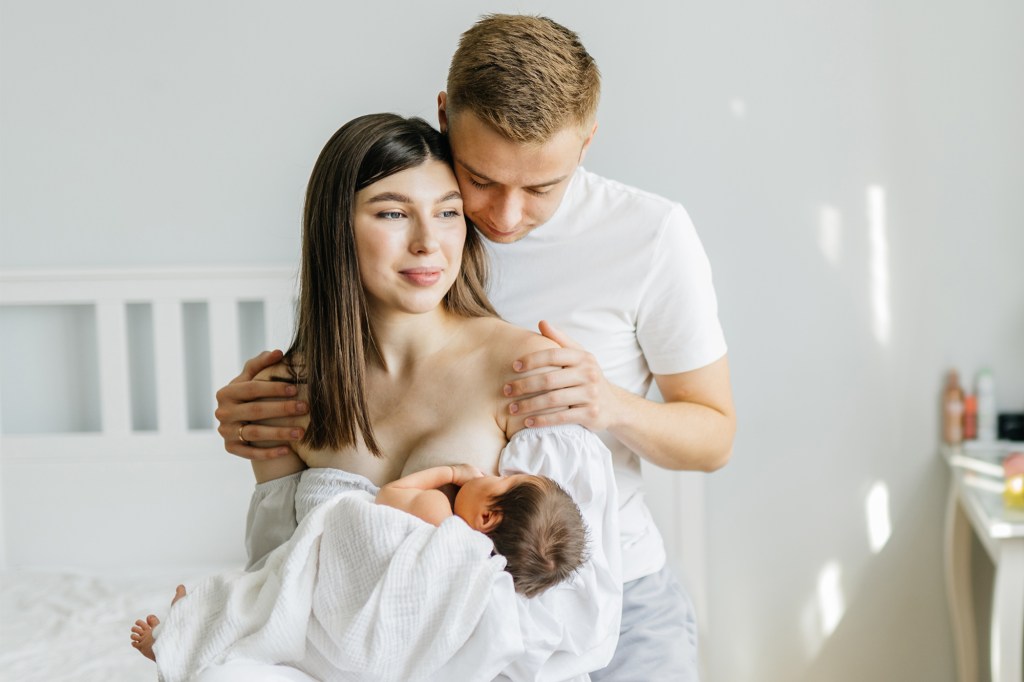 Couple in love with similar Mario Novembre, sitting in a cozy bedroom; the woman breastfeeding her newborn daughter while the man gives her a warm hug.